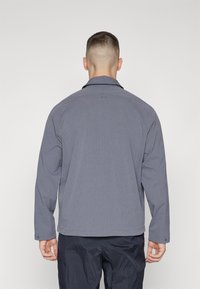 Gray, lightweight jacket with a subtle texture, long sleeves, and a collar. Features buttoned cuffs and a logo on the back.