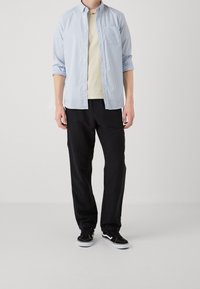 Chemise à rayures bleues et blanches à boutons, portée sur un t-shirt beige, associée à un pantalon noir et des baskets noires. Tissu uni avec un design simple.