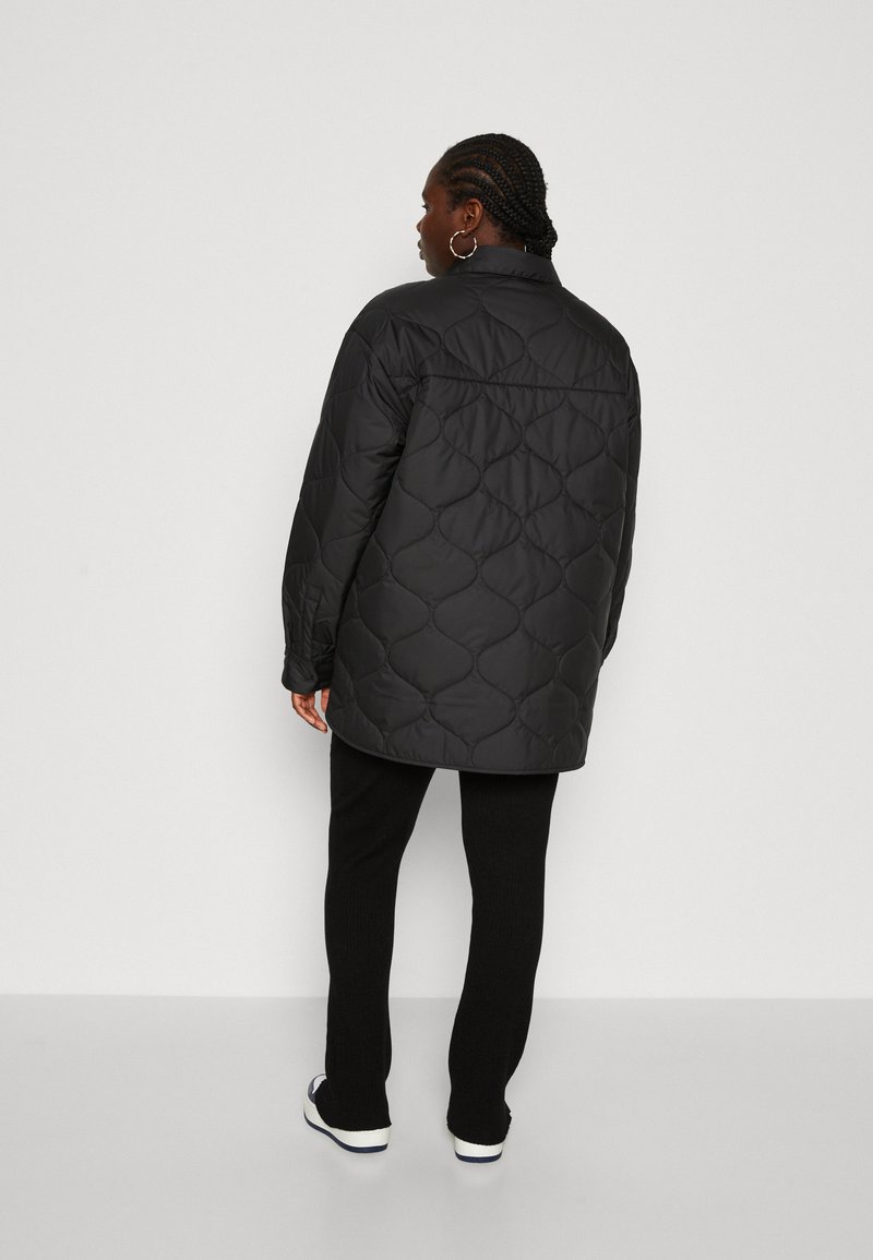 Manteau long matelassé noir avec col montant, présentant un motif géométrique et une coupe décontractée. Assorti à un pantalon noir et des baskets blanches.