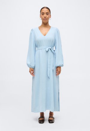 Kvinne står iført en lys blå langermet maksimalang kjole med belte knyttet i midjen og svarte flate sko med stropper.