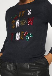 Pull en tricot bleu marine avec le texte en sequins multicolores "C'EST L'HEURE DU SPECTACLE" et des accents de houx. Présente un col et un ourlet côtelés.