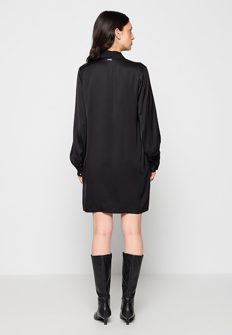 Abito in seta nera con maniche lunghe, colletto e leggero lucido. L'abito ha una vestibilità comoda, raggiunge la metà della coscia ed è abbinato a stivali alti fino al ginocchio.