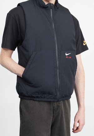 Osoba ubrana w czarną bezrękawnik z zamkiem i logo Nike Air, czarną koszulkę z krótkim rękawem oraz ciemne spodnie, jedna ręka w kieszeni kamizelki.