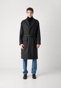 Dark gray, knee-length wool blend coat with a wide lapel, belt at the waist, two front pockets, worn over a black turtleneck and blue jeans.