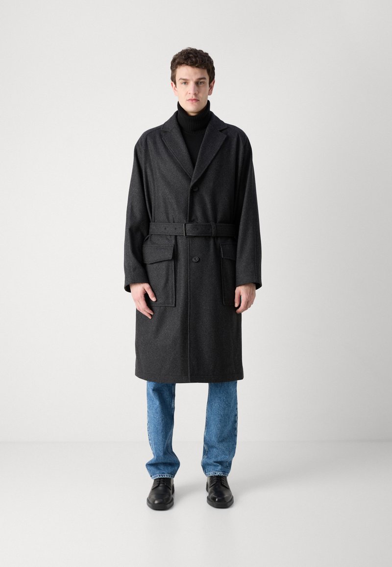 Manteau en laine mélangée gris foncé, longueur genou, avec un large revers, une ceinture à la taille, deux poches avant, porté sur un col roulé noir et un jean bleu.