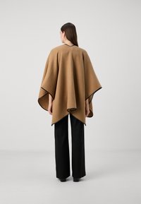 Brun uldblandings-poncho med et flydende design og sorte detaljer, parret med sorte brede bukser. Teksturen er blød og varm.