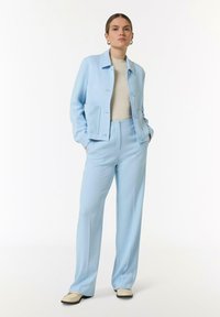 Veste ajustée bleu clair avec une seule poche et des boutons, associée à un pantalon plissé assorti, portée sur un pull beige.