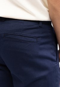 Pantaloni blu navy realizzati in tessuto morbido, con una tasca posteriore dotata di un flap rettangolare. Il tessuto ha una leggera lucentezza ed è ben tagliato.