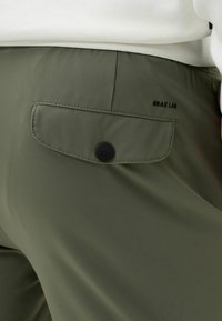 Olivově zelené kalhoty s kapsou na zadní straně, která má zapínání na knoflík a embosované logo. Materiál vypadá hladce a má matný povrch.