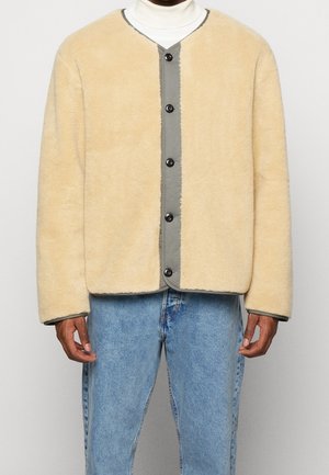 Man wearing beige buttoned fleece jacket over white turtleneck and light blue denim jeans, standing against plain background.