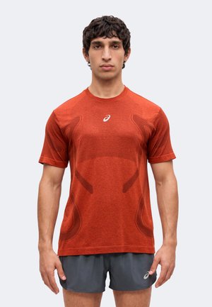 Junger Mann mit dunklem lockigem Haar, der ein oranges kurzärmliges Sportshirt und dunkelgraue Laufshorts trägt und nach vorne schaut.