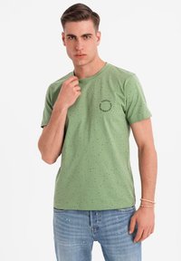 Camiseta de algodón verde con cuello redondo, mangas cortas y un patrón salpicado de manchas negras. Pequeño estampado negro en el pecho.