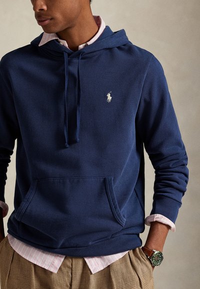 Bărbat purtând un hanorac bleumarin peste o cămașă roz cu dungi, pantaloni bej în carouri și un ceas verde cu curea maro din piele, mâinile în buzunare.