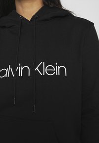 Felpa nera con cappuccio realizzata in morbido tessuto, dotata di tasca frontale, cappuccio regolabile e grande scritta bianca "Calvin Klein" sulla parte anteriore.