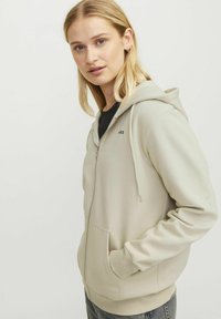 Hoodie beige zippé avec une poche avant, capuche ajustable et poignets côtelés. Présente un logo subtil sur la poitrine. Texture de tissu lisse.