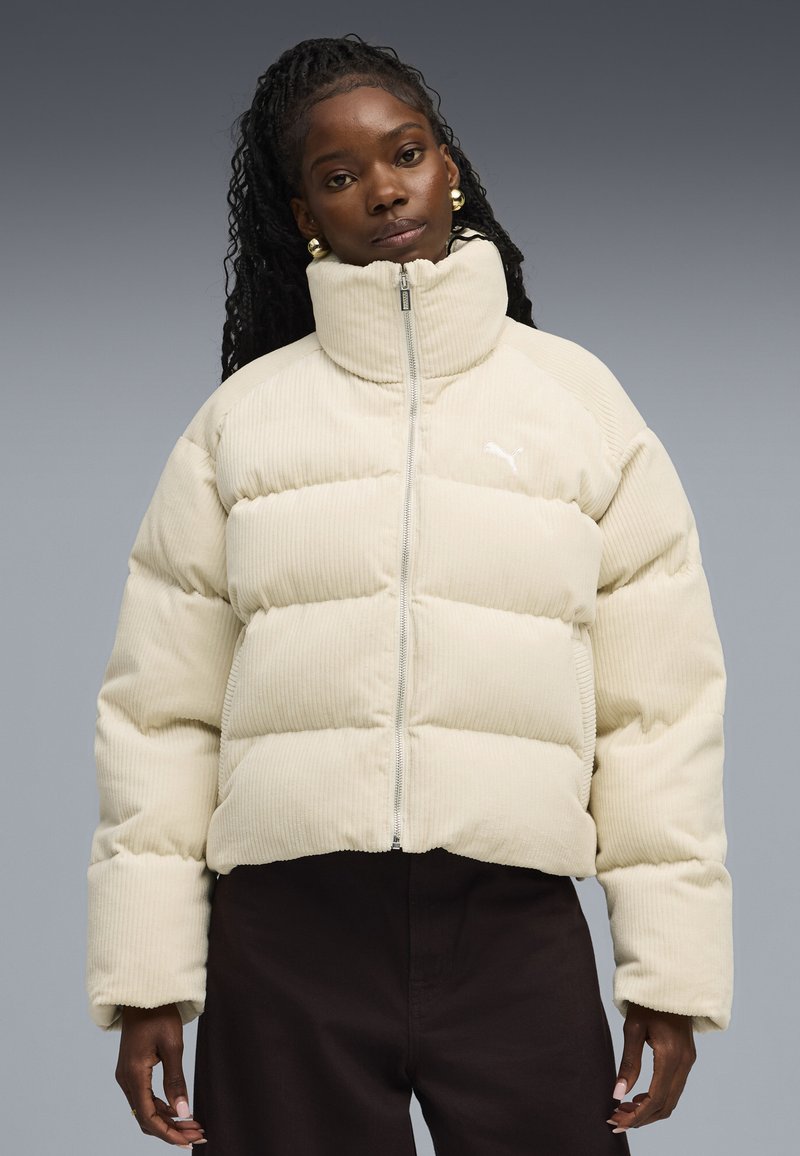 Beige corduroy pufferjack met hoge kraag en voorritssluiting. Bevat gewatteerde secties en een klein logo aan de linkerkant.