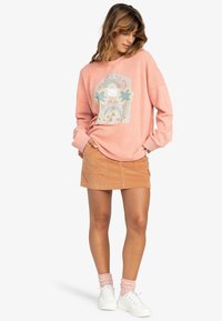 Rosa übergroßes Sweatshirt mit einem Blumenmotiv, kombiniert mit einem beigefarbenen Cordrock. Das Model trägt weiße Sneakers und rosa strukturierte Socken.