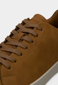 Gros plan d'une basket en daim marron avec des lacets plats assortis et des coutures visibles sur la tige, posée sur un fond blanc.