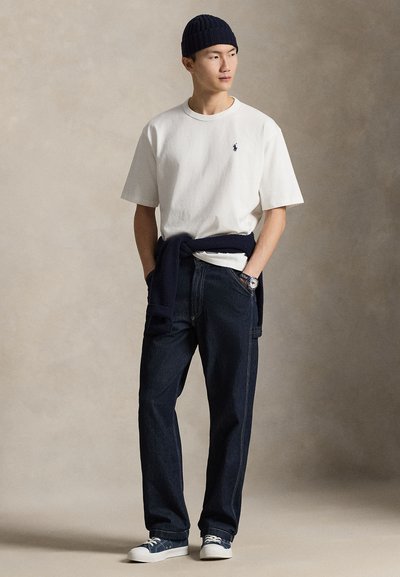 T-shirt en coton blanc avec logo, bonnet en maille bleu marine, pantalon en jean foncé et baskets blanches. Style complété par un pull bleu marine noué autour de la taille.