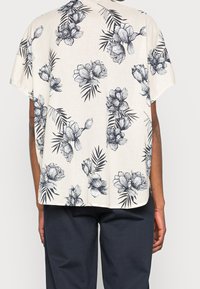 Chemise imprimée florale en crème avec des fleurs et des feuilles bleu foncé. Coupe ample, manches courtes et ourlet droit. Assortie à un pantalon foncé.