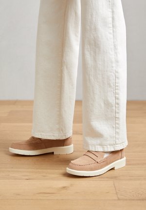 Suède instapschoenen in licht beige met een witte zool, voorzien van decoratieve stiksels en een taps toelopend ontwerp. Gecombineerd met crèmekleurige broek met wijde pijpen.