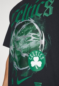 Sort t-shirt med et grønt og sølv grafisk design, der viser "Boston Celtics" og et kløverlogo. Stof har en glat tekstur.