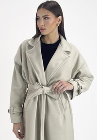 Cappotto trench beige chiaro con cintura realizzato in tessuto testurizzato. Caratterizzato da ampie rever, tasche laterali e maniche arrotolate con dettagli a fibbia.