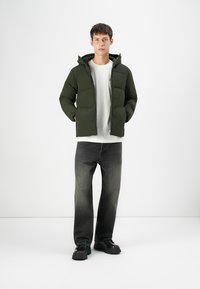 Dark green padded jacket with a hood, zip front, and side pockets, paired with a white t-shirt and gray jeans, worn with black shoes.
