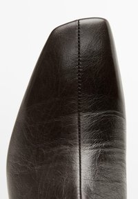 Chaussure en cuir noir avec un bout pointu, présentant une texture lisse et des coutures détaillées le long de la couture. Design minimaliste, sans embellissements.