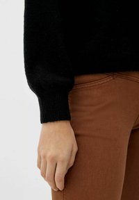 Linke Hand ruht neben der Hosentasche der braunen Hose, trägt einen schwarzen Pullover mit langen Ärmeln und gerippten Bündchen.