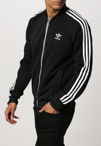 Man wearing black Adidas track jacket with white stripes on sleeves and logo on chest, hands partially in pockets.