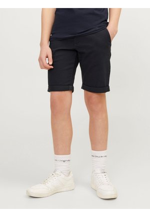 Personne portant un short noir retroussé, des chaussettes blanches avec texte noir et des baskets blanches, debout devant un fond clair uni.