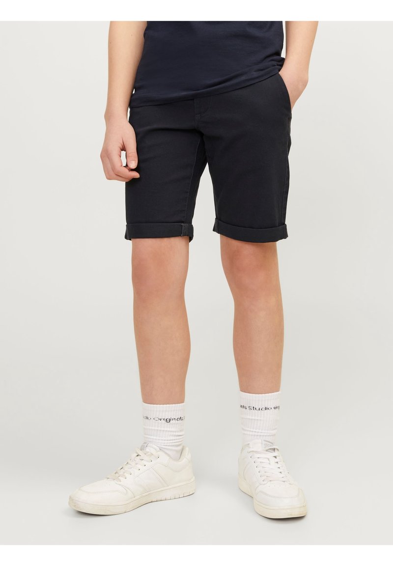 Personne portant un short noir retroussé, des chaussettes blanches avec texte noir et des baskets blanches, debout devant un fond clair uni.