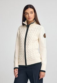 Krämfärgad kabelstickad tröja med hög krage, grön dragkedja, kontrasterande ribbade muddar och nederkant samt en broderad logotyp på ärmen.