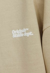 Béžová mikina vyrobená z měkké tkaniny, s bílým vyšitým textem "Originals™ Studio dept."