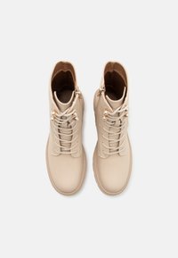 Beige lærankelsko med rund tå, snørebånd foran og glidelås på siden. Har gullfarget detaljer og en glatt tekstur.