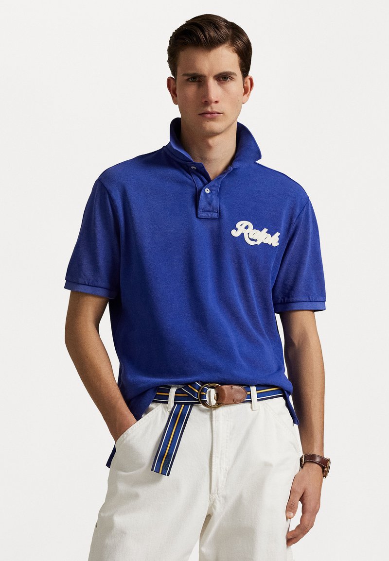 Tricou polo albastru din bumbac, cu guler și logo alb brodat. Asortat cu pantaloni albi și o curea cu dungi multicolore.