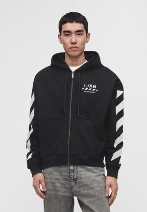 Jeune homme portant un sweat à capuche noir zippé avec des bandes diagonales blanches sur les manches et un texte graphique blanc sur la poitrine, associé à un jean gris.