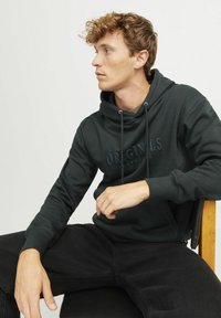 Dark green hoodie with drawstrings, featuring "ORIGINALS" logo in text on chest, paired with black pants, seated on a chair.