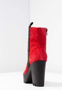 Botín de tobillo de ante rojo con grueso tacón negro de plataforma, con una tiradora negra que presenta el texto "Calvin Klein Jeans" y cremallera lateral.
