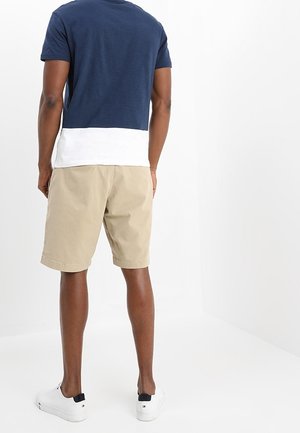 Homme debout, de dos, portant un t-shirt bleu marine et blanc, un short beige et des baskets blanches sur un fond blanc.