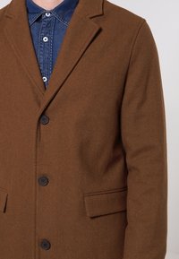 Cappotto in misto lana marrone con rever a scollo a V, design monopetto, tre bottoni neri e due tasche laterali. Indossato sopra una camicia di denim.