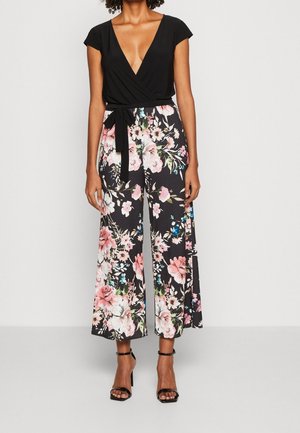 Femme portant un haut noir cache-cœur à col en V profond et un pantalon large noir avec un imprimé floral rose ainsi que des sandales noires à talons hauts.
