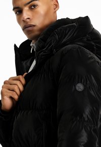 Chaqueta de plumas negra con acabado brillante, que presenta un cuello alto, un acolchado grueso y un parche redondo con logo en la manga izquierda. Cierre con cremallera visible.