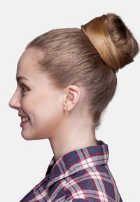 Brunt hår stylat i en knut, bärande en guldsmyckeshängare. Klädd i en röd och blå rutigt skjorta. Profilbild mot en enfärgad bakgrund.