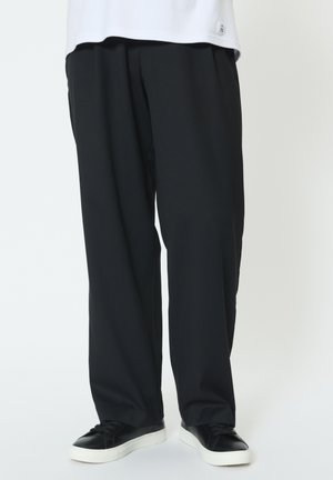 Man wearing loose black trousers and black sneakers with white soles standing against a white background.