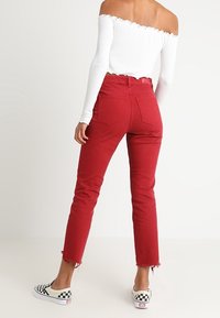 Jean en denim rouge avec un ourlet effiloché, taille haute et poches arrière, assorti à un haut blanc aux épaules dénudées et des chaussures sans lacets à carreaux noir et blanc.