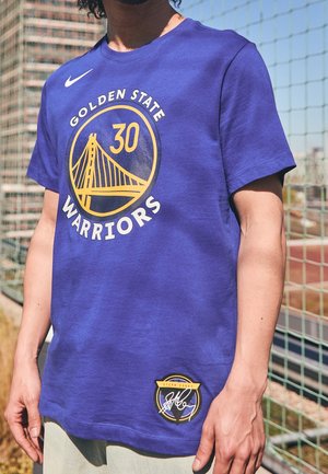 Osoba ubrana w niebieską koszulkę Golden State Warriors z numerem 30 i logo mostu, stojąca przy ogrodzeniu w słoneczny dzień.