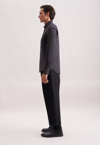 Camicia grigia a maniche lunghe con un motivo testurizzato, abbinata a pantaloni neri e scarpe. La camicia ha un orlo curvo e polsini con bottoni.