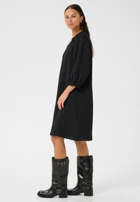Robe noire, texturée, longueur genoux avec des manches bouffantes et un décolleté en V, associée à des bottes en cuir noir poli à talons avec des boucles.